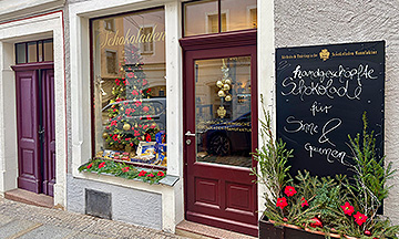 Ein Geschäft mit einer roten Tür und einem großen Fenster zeigt einen geschmückten Weihnachtsbaum, Schmuck und festliche Girlanden. Draußen steht eine Tafel mit der Aufschrift "Handgeschöpfte Schokolade für Sinne & Gaumen", an deren Fuß rote Blumen angebracht sind.
